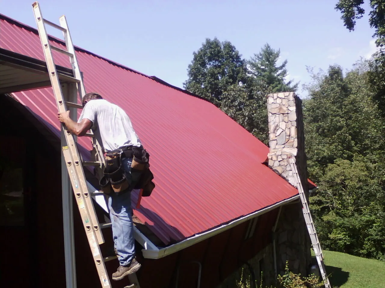 Roofer on a ladder beside a red metal roof and stone chimney — Soffit & Fascia Repair by Roofing Brothers in Sardis